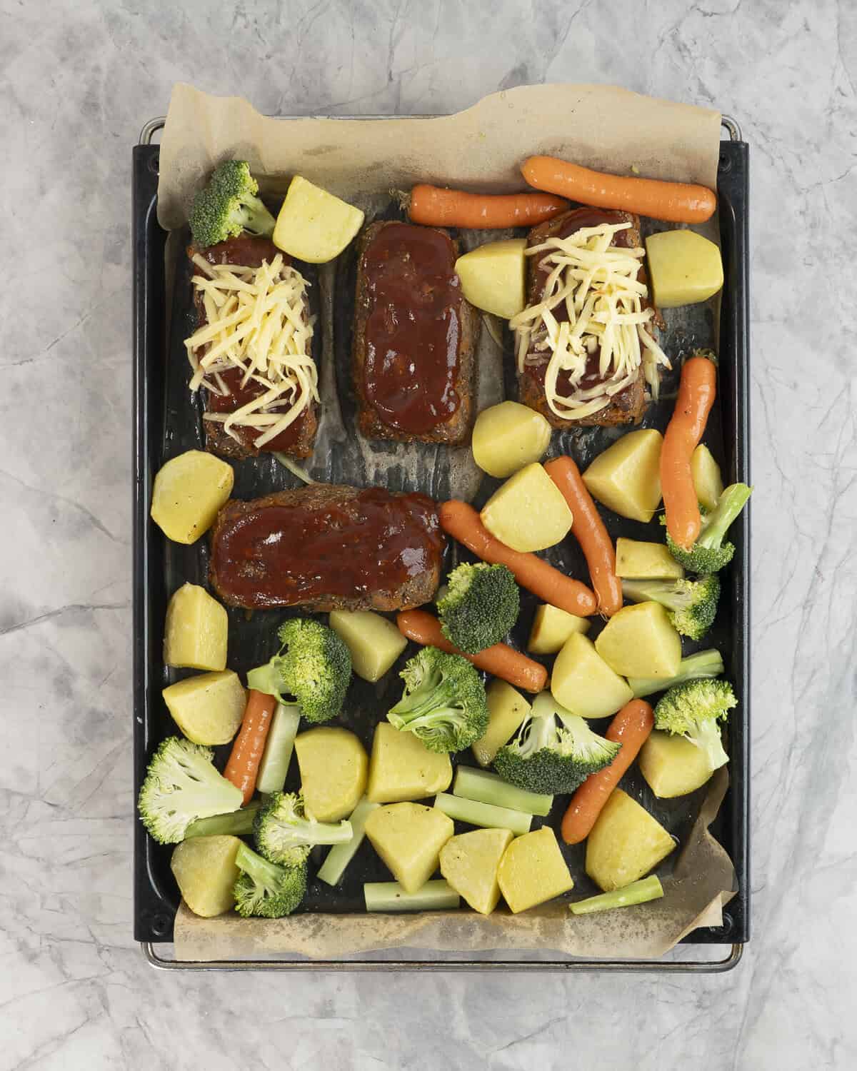 Lined baking tray with chopped potato, broccoli, mini carrots and four mini rectangle meatloaf's two topped with tomato sauce and grated cheese and 2 with tomato sauce.