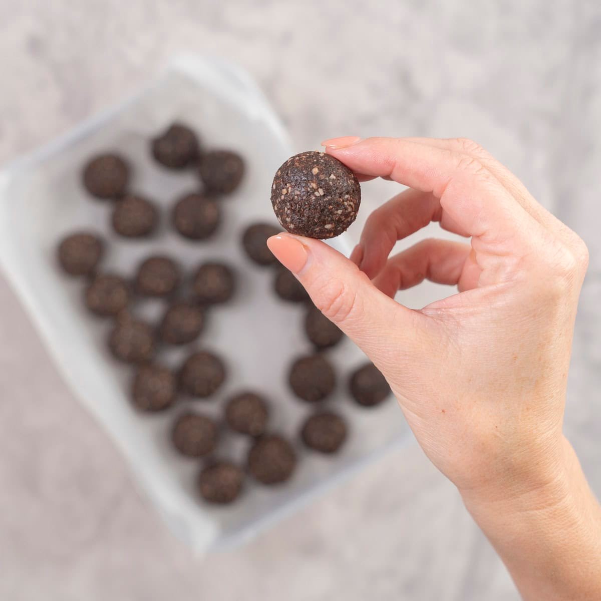 Womans hand holding a small round weetbix bliss ball up to camera, container of other balls in background on benchtop.