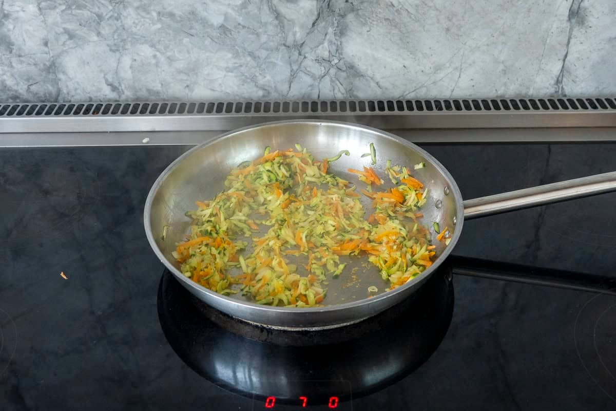 grated onion, zucchini and carrot cooking in a stainless steel skillet.