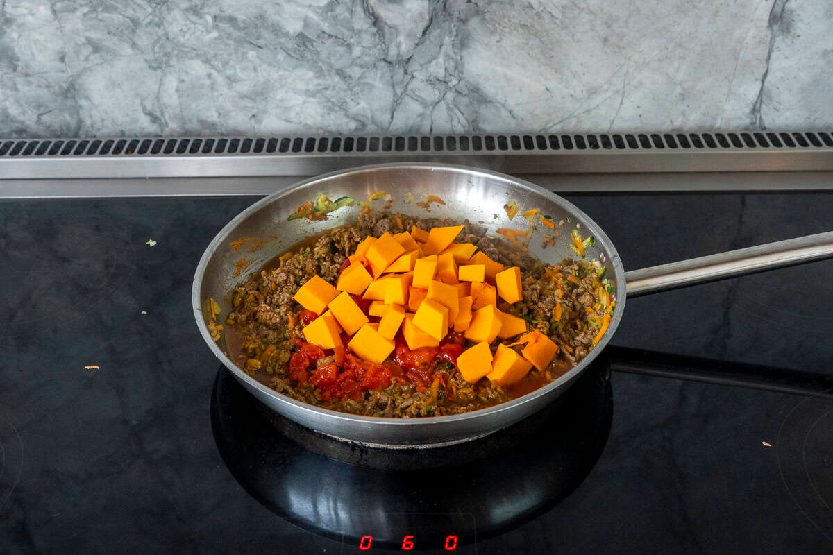 cubes of pumpkin and tinned tomatoes added on top of a skillet of browned mince and vegetables.