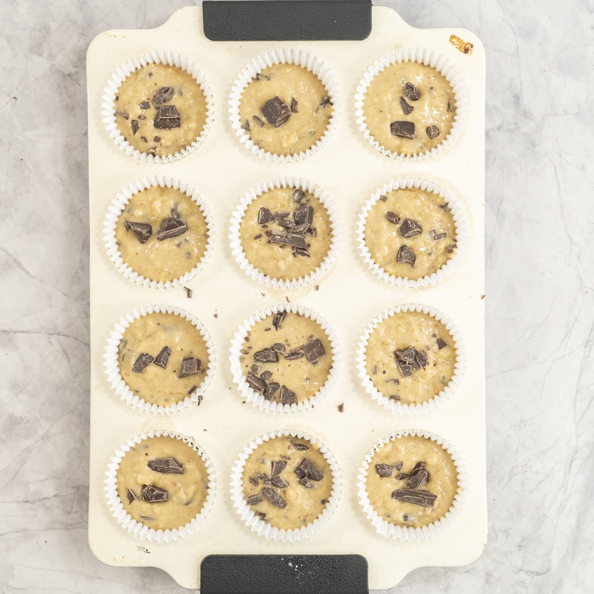 Muffin tray filled with banana chocolate chunk muffin batter inside and chocolate sprinkled on top.