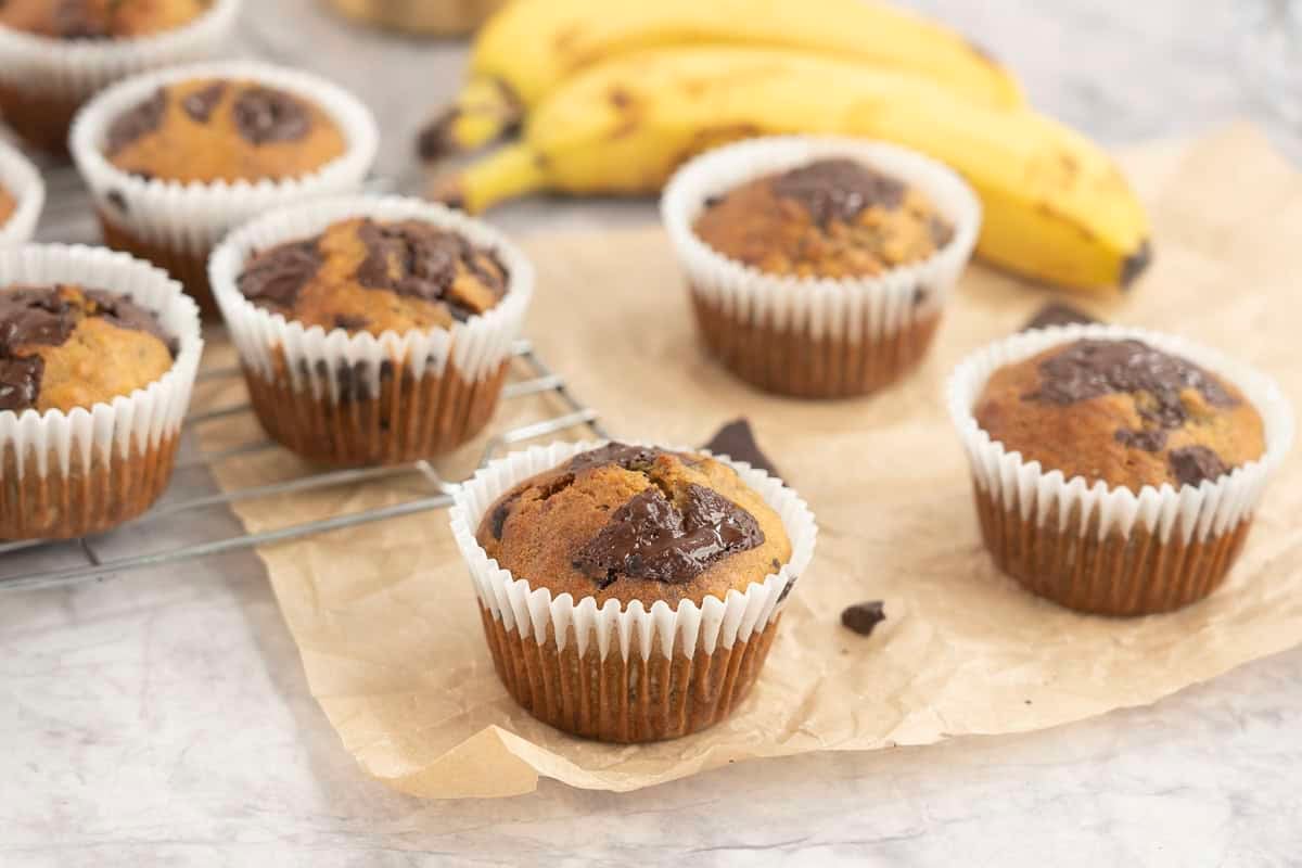 Cooked banana chocolate chunk muffins on cooling rack and baking paper on bench top with gooey melted chocolate on top.