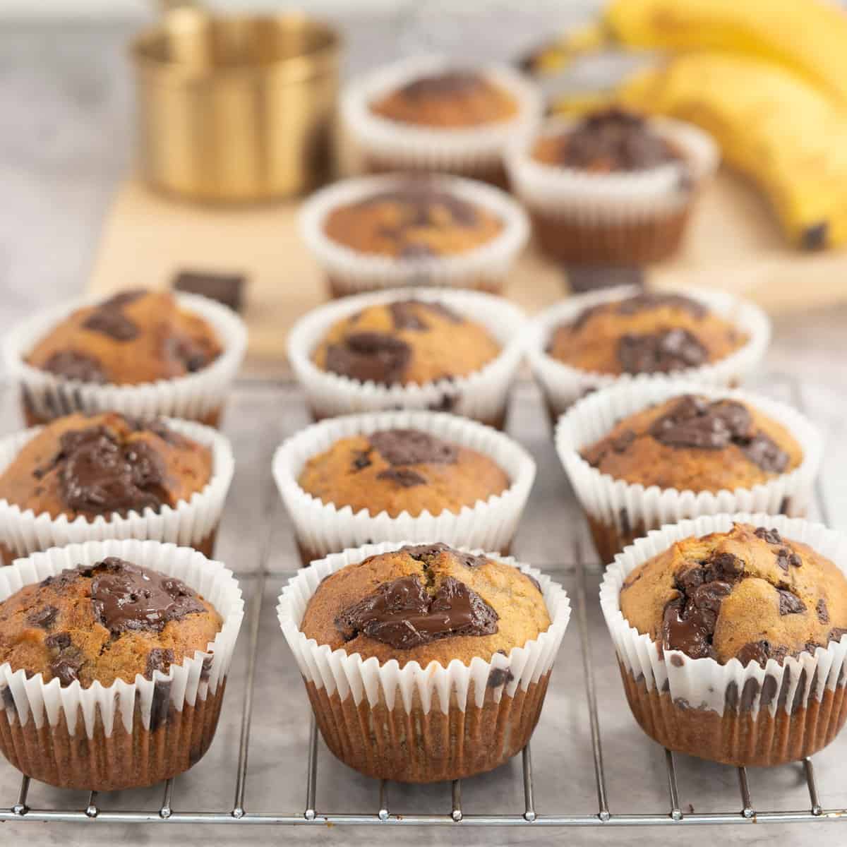 Cooked banana chocolate chunk muffins on cooling rack and baking paper on bench top with gooey melted chocolate on top.