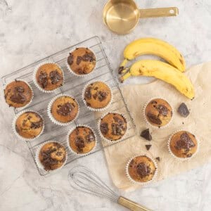 Cooked banana chocolate chunk muffins on cooling rack and baking paper on bench top with gooey melted chocolate on top.