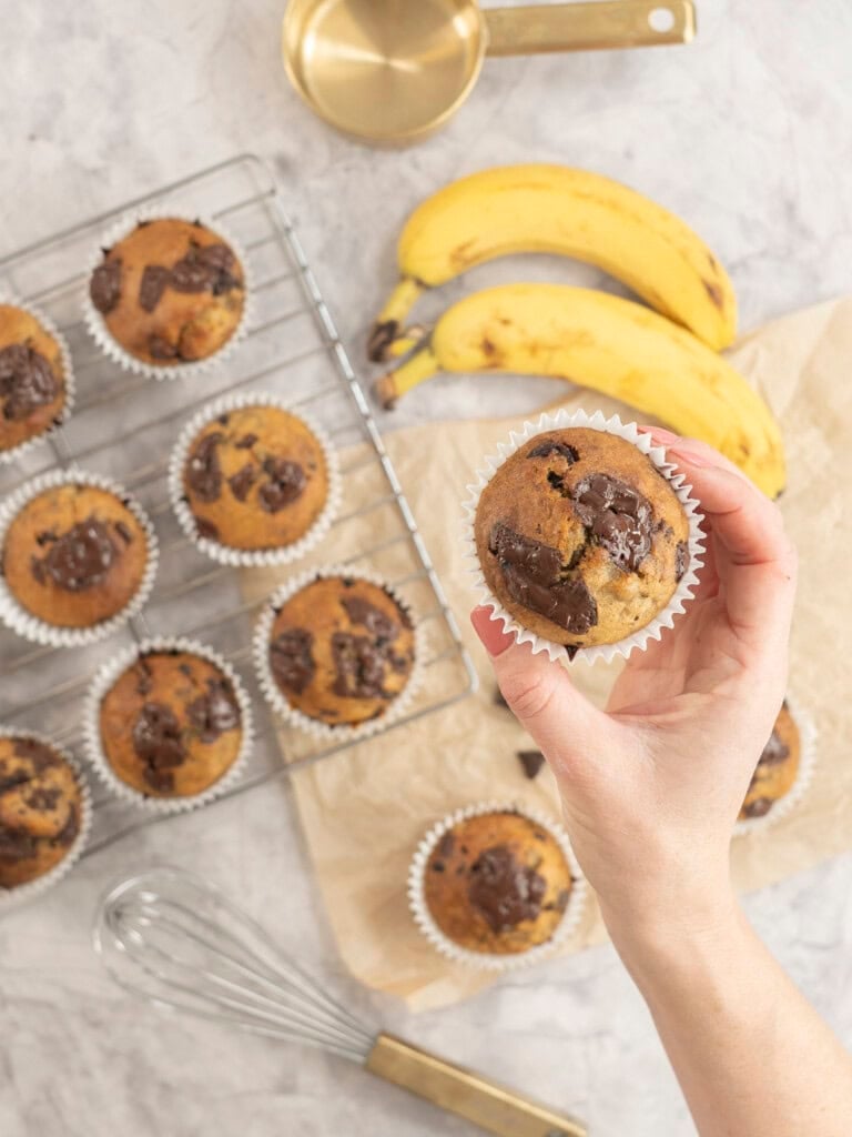 Hand holding a cooked banana chocolate chunk muffin, more below on cooling rack and baking paper on bench top with gooey melted chocolate on top.