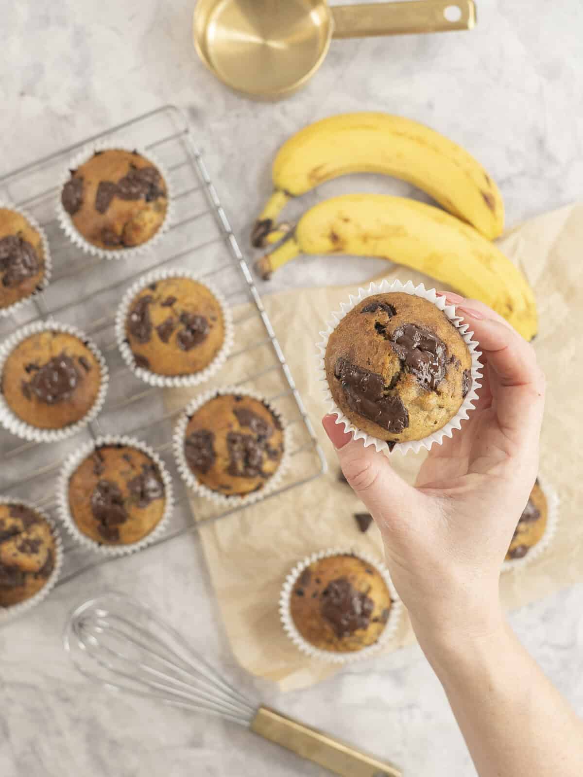 Hand holding a cooked banana chocolate chunk muffin, more below on cooling rack and baking paper on bench top with gooey melted chocolate on top.