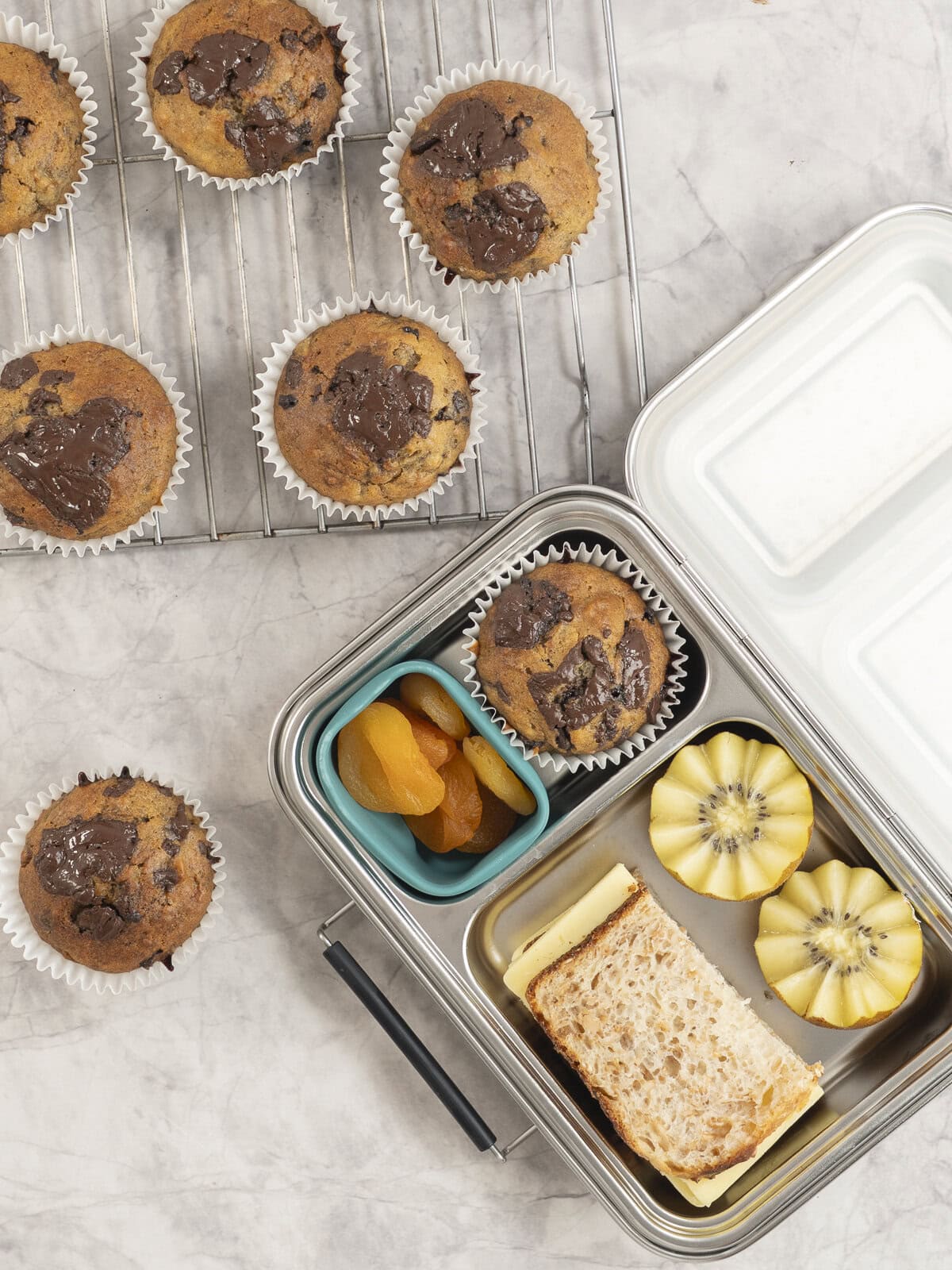 An open lunchbox on benchtop with a cheese sandwich, halved kiwifruit, dried apricots and chocolate chunk muffin inside. Muffins on cooling rack on benchtop.