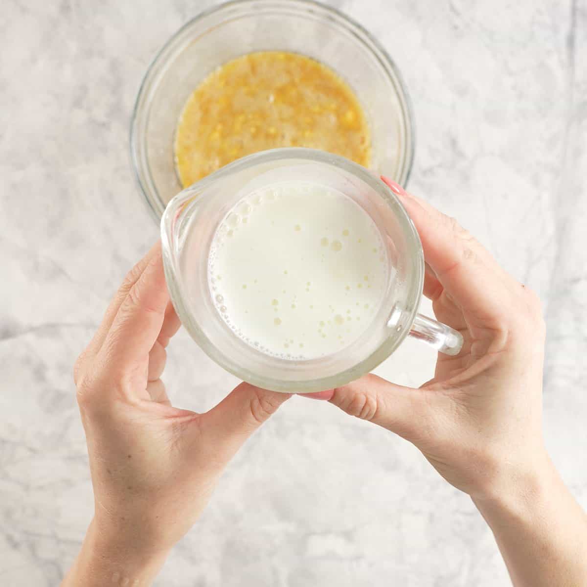 Hands holding a glass jug of milk above wet mixture on benchtop.