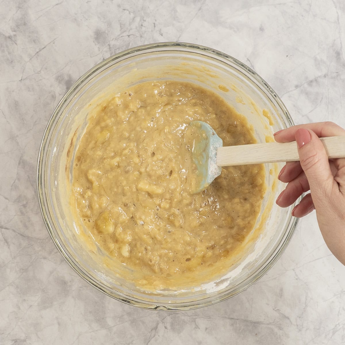 Wet and dry ingredients mixed together with hand holding a spatula in glass bowl on bench top.
