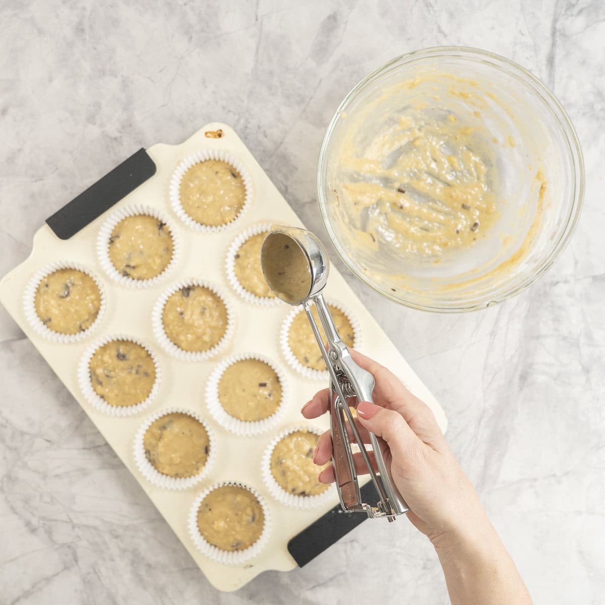 Hand holding an ice cream scoop portioning muffin batter into the muffin trays.