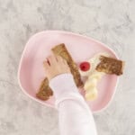 A baby's hand reaching towards a plate of French toast berries and yogurt.