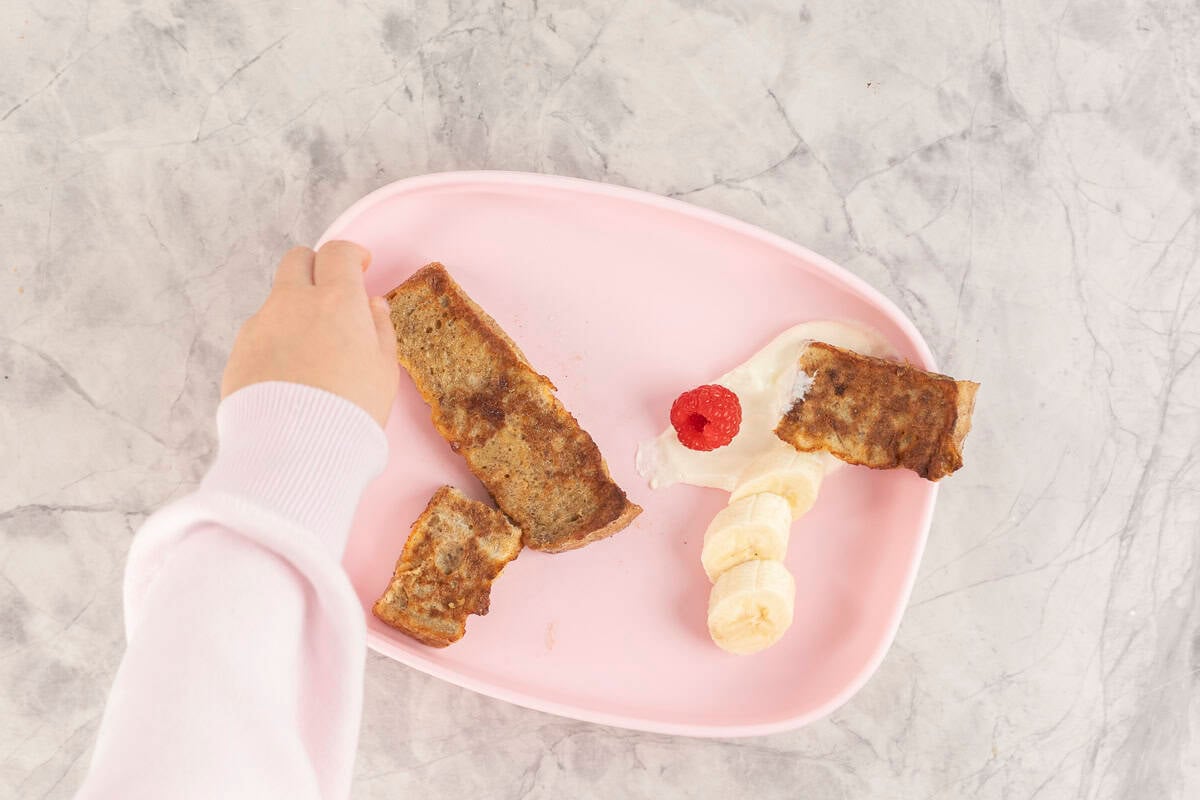 a baby picking up a strip of toast from a pink baby plate of toast, yogurt and raspberries.