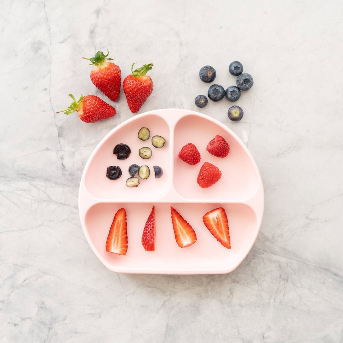 A pink silicone divided baby plate with different types of berries prepared for baby led weaning.