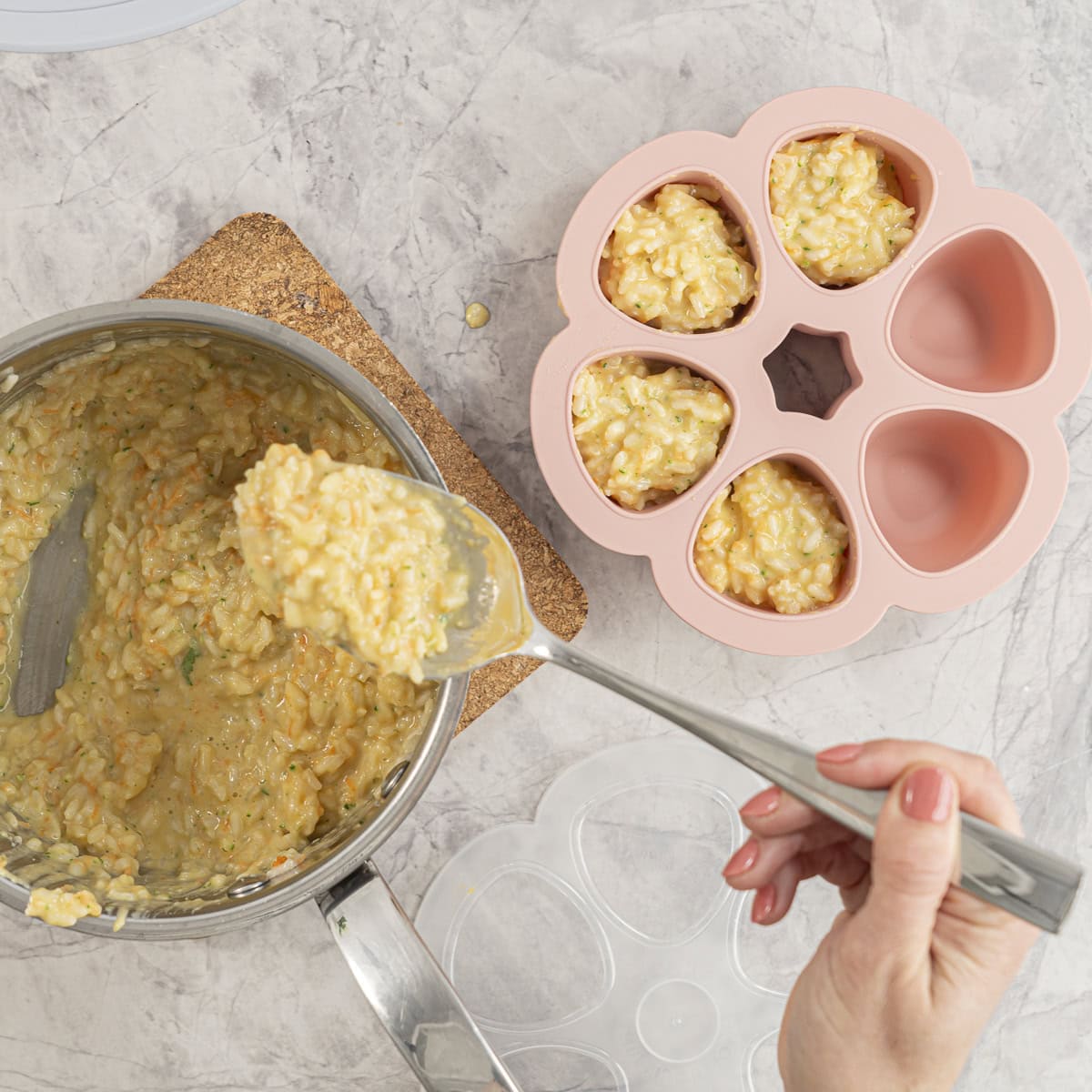 Pot on cork board on bench top with hand holding a serving spoon with scoop of creamy rice on it, pink silicone mould of six sections with cheesy rice inside.