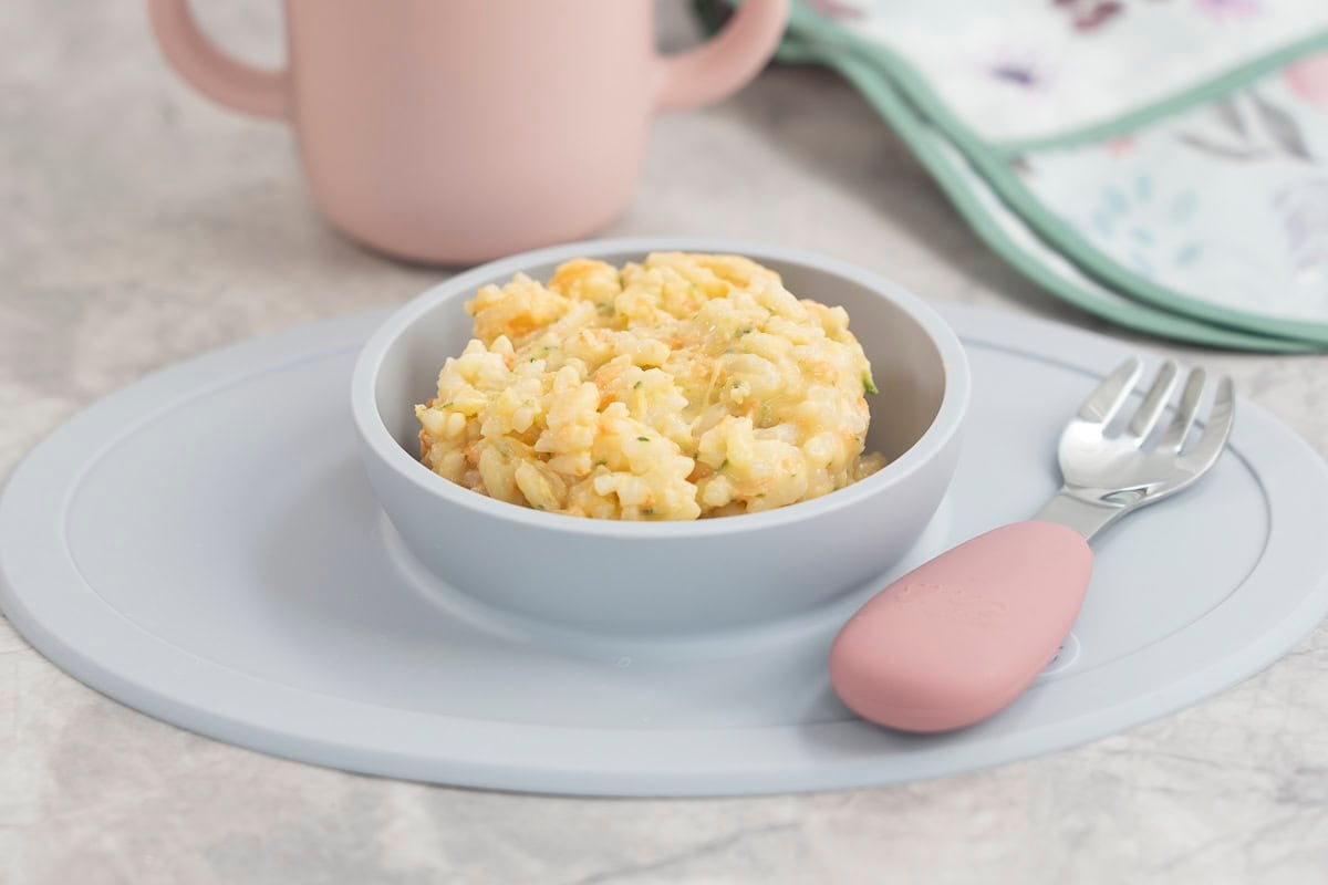Blue silicone baby bowl with cheesy rice inside, toddler fork with pink silicone handle, pink silicone babu cup on benchtop.