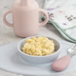 Blue silicone baby bowl with cheesy rice inside, toddler fork with pink silicone handle, pink silicone babu cup on benchtop.