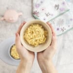 Woman holding a bowl with cheesy rice inside above a silicone baby plate, drink bottle and bib below on bench top.