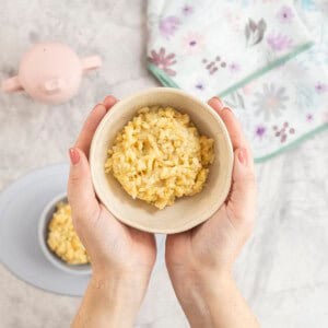 Woman holding a bowl with cheesy rice inside above a silicone baby plate, drink bottle and bib below on bench top.