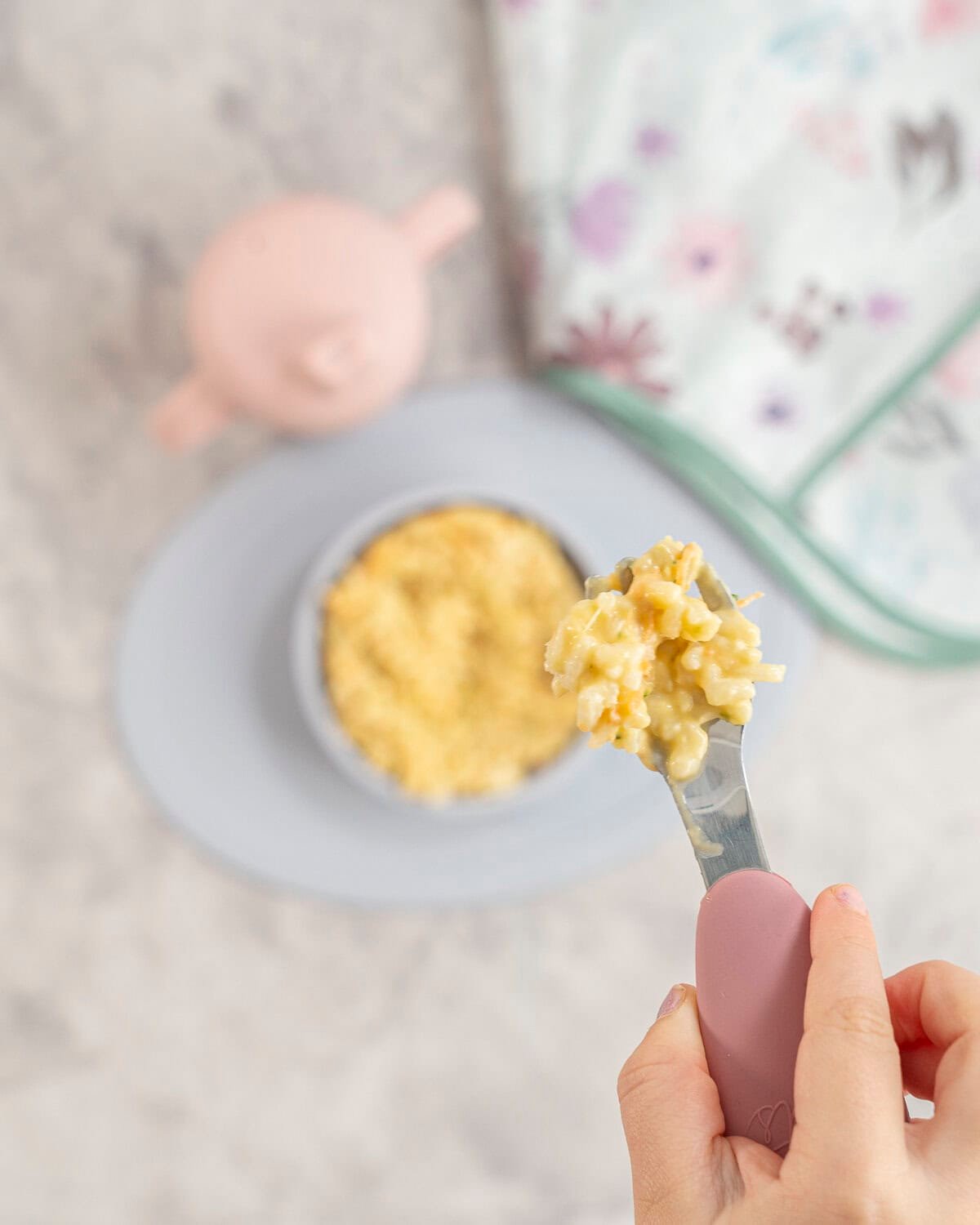 Toddlers hand holding a pink handled baby fork with a scoop of cheese rice on it taken from silicone baby bowl below on bench top with cheesy rice inside.
