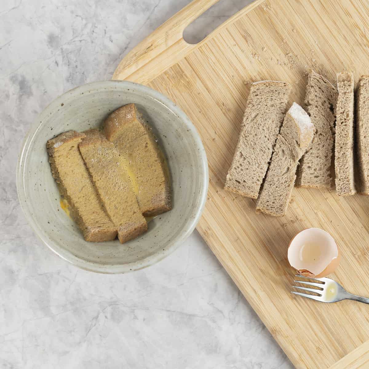 Bread cut into three slices in a bowl of whisked egg next to a wooden chipping board with sliced bread on it.