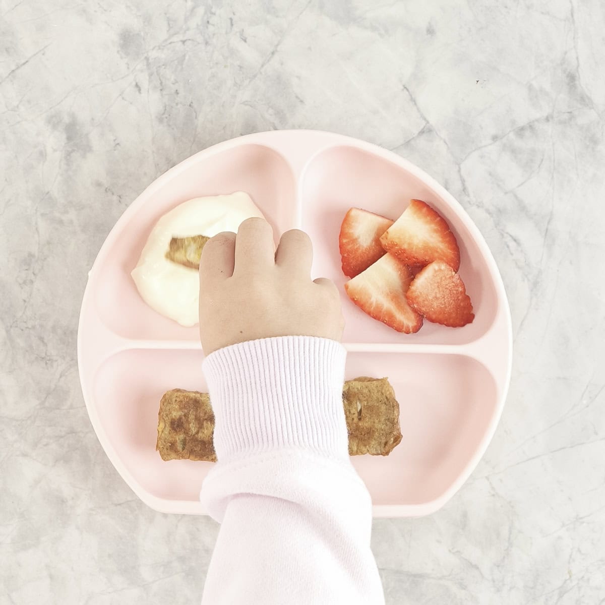 Toddler hand holding a slice of eggy bread dipping it into yogurt in a pink silicone plate with cut strawberries inside.