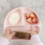 A toddlers hand taking a piece of eggy bread from a pink silicone baby plate with cut strawberries and yogurt.