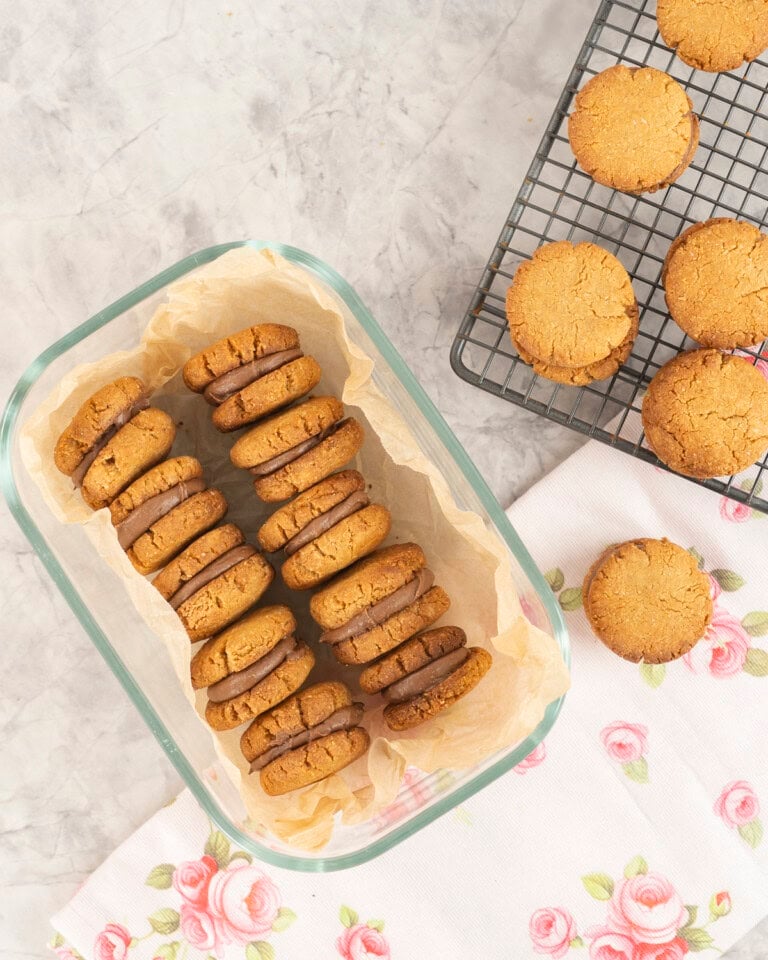 Glass container with two rows of peanut butter sandwich cookies inside next to cooling tray with more cookies on top.