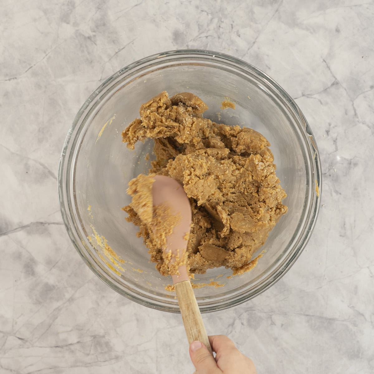 Cookie dough being mixed with a spatula in a large glass bowl on bench top.