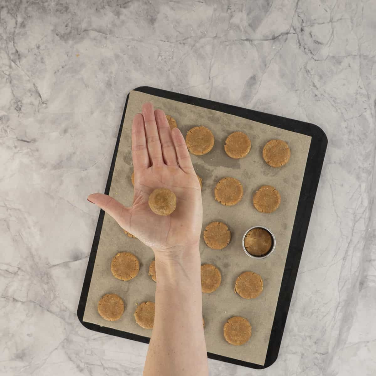 Woman holding a small ball of cookie dough in her palm, lined baking tray with small round flat cookies.