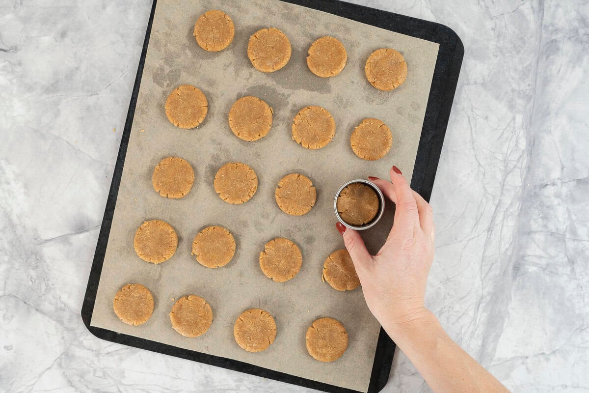 Woman holding a small circle cookie cutter around cookie on lined baking tray.