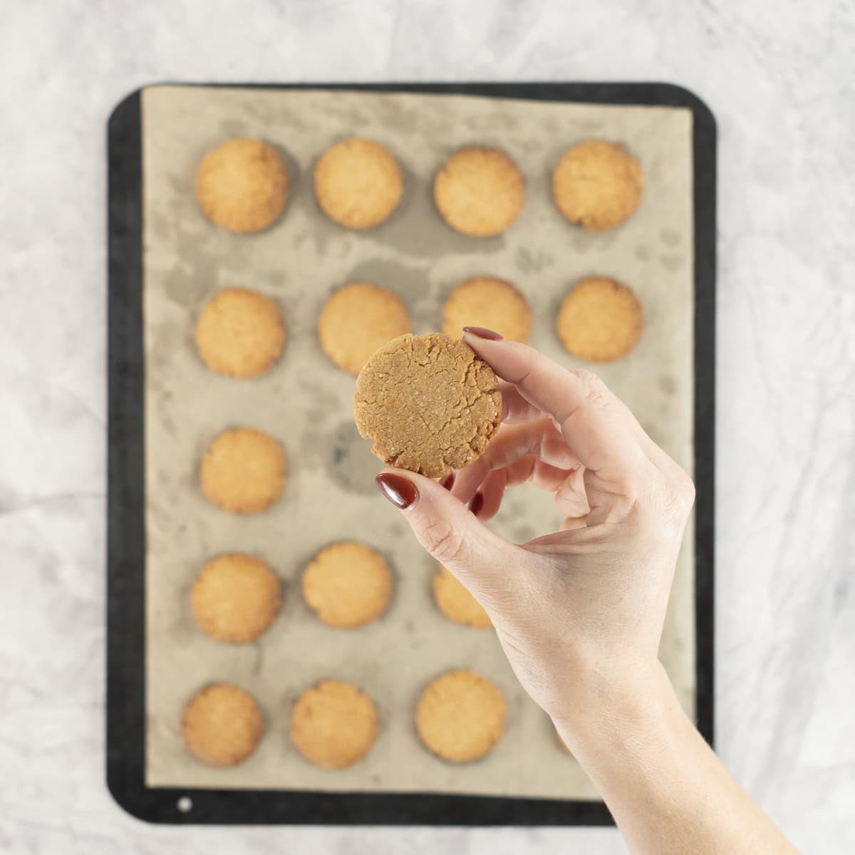 Hand holding a small cookie above lined baking tray with batch of cookies.