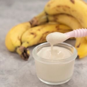 Small glass container with pink baby spoon scooping out banana pudding, bananas stacked in background.