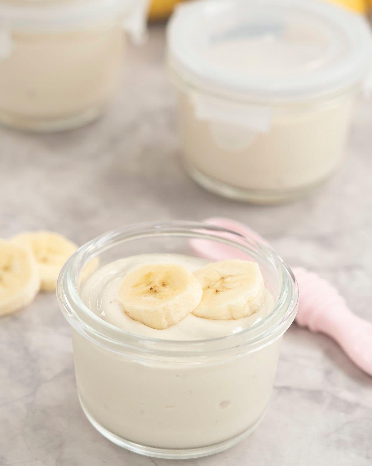 Four small glass containers with banana pudding inside, one with no lid on and two slices of banana on top and a pink baby silicone spoon.