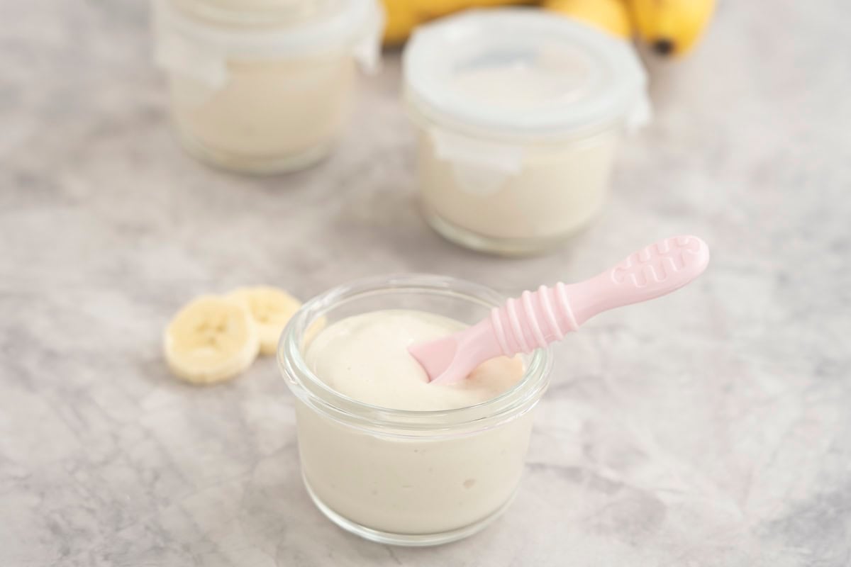 Four small glass containers with banana pudding inside, one with no lid on and a pink baby silicone spoon inside.