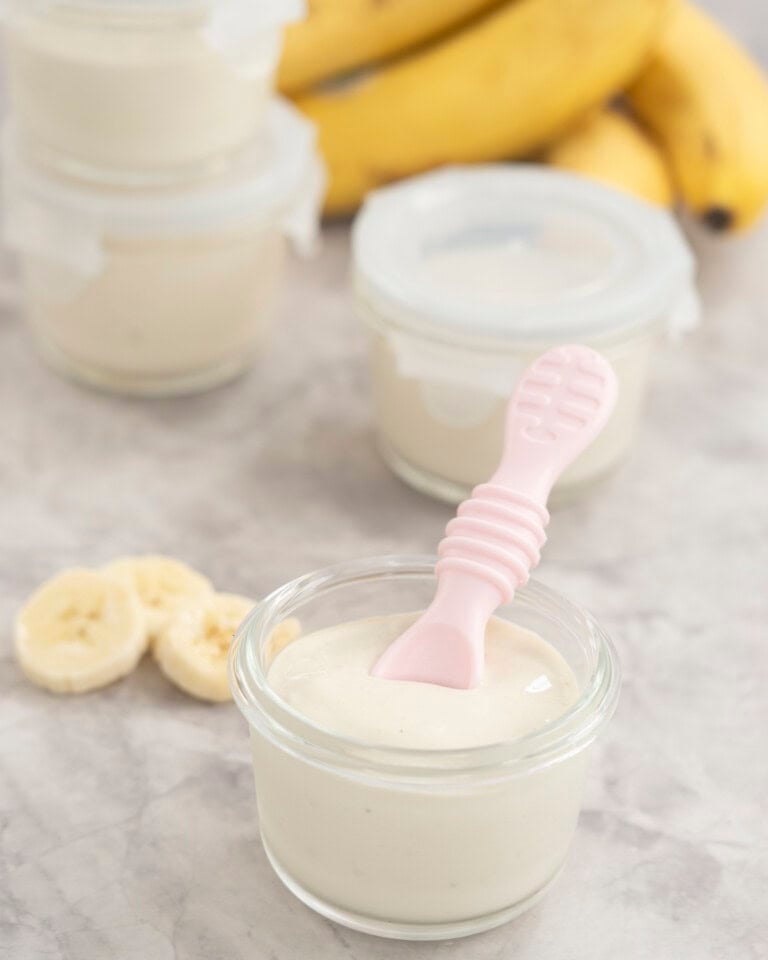 Four small glass containers with banana pudding inside, one with no lid on and a pink baby silicone spoon inside.