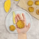 A Childs hand holding a meatball above a baby plate of fruit and vegetables.