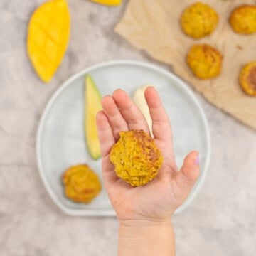 A Childs hand holding a meatball above a baby plate of fruit and vegetables.