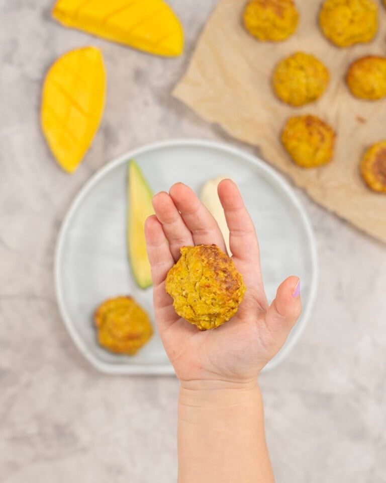 A Childs hand holding a meatball above a baby plate of fruit and vegetables.