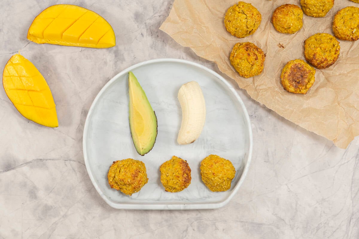 Three mango chicken meatballs on a baby plate with a piece of avocado and banana, next to a mango cut hedgehog style.