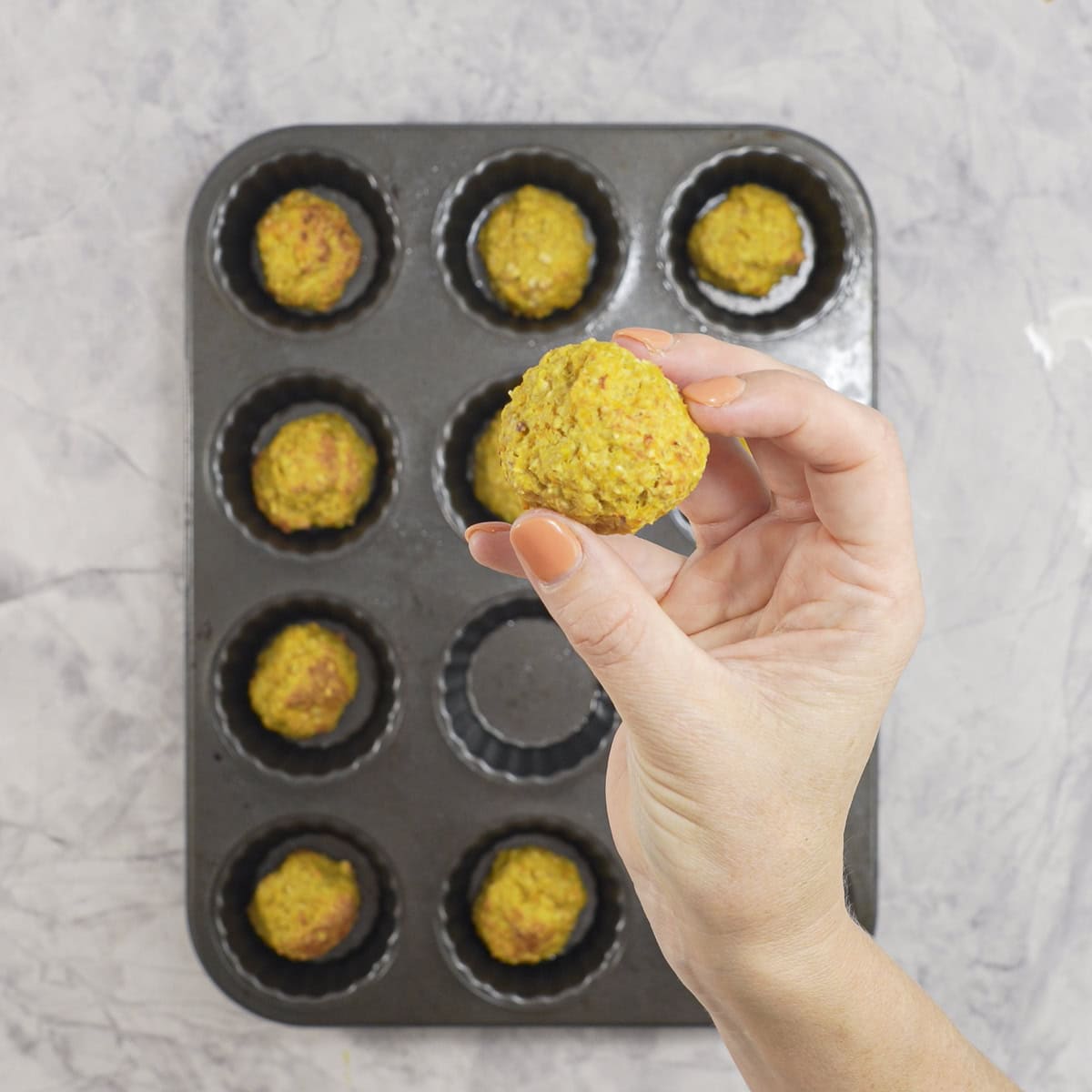 A golden baked meatball being held above a muffin tray of baked meatballs.