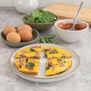Pizza eggs sliced into four pieces on a plate, serving dishes in background with spinach, tomato paste and eggs.