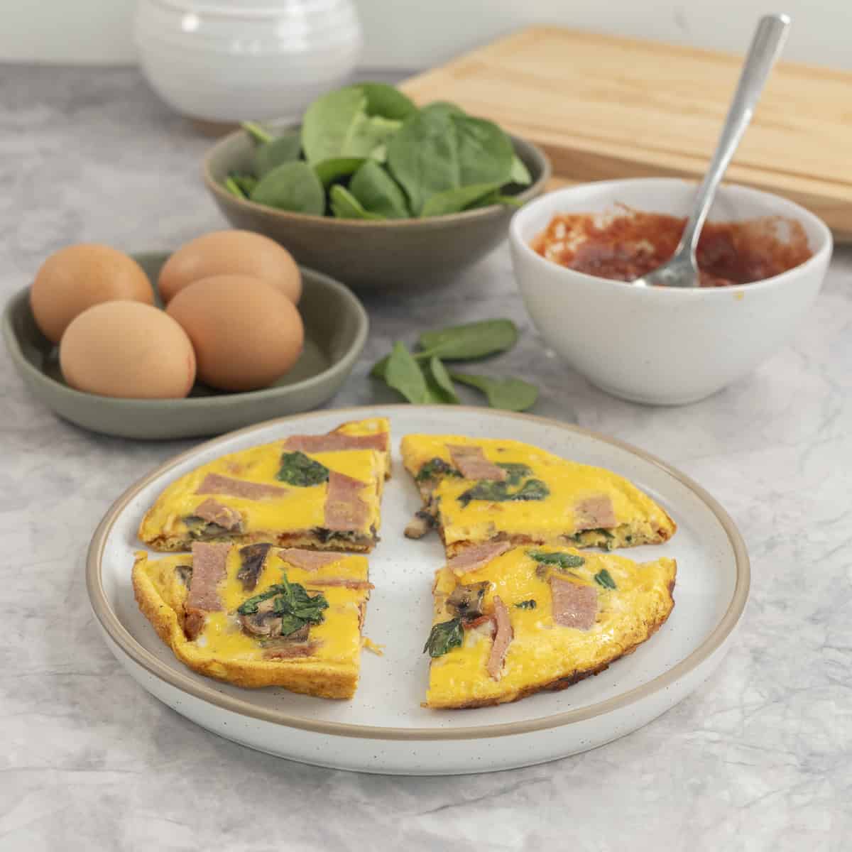 Pizza eggs sliced into four pieces on a plate, serving dishes in background with spinach, tomato paste and eggs.