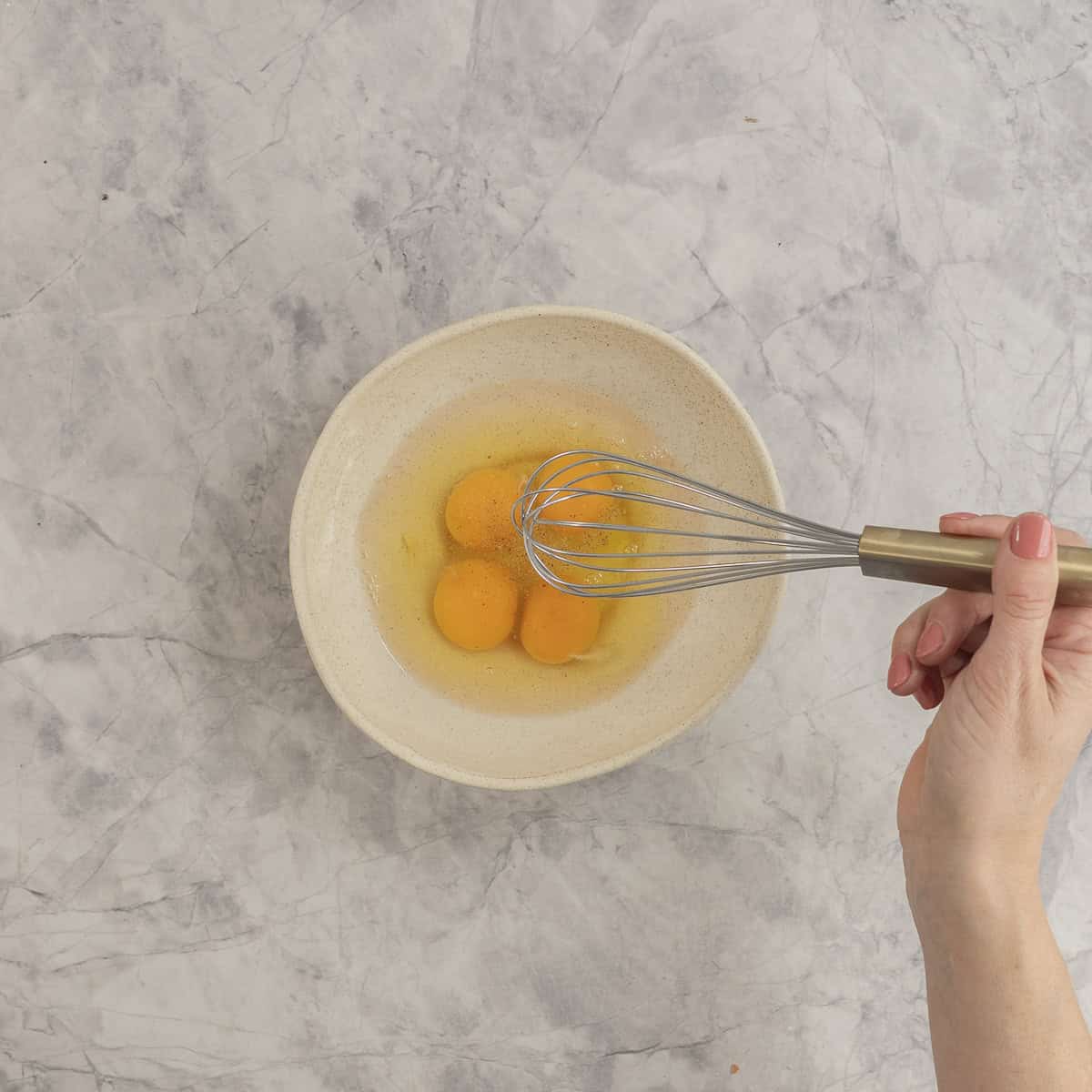 Womans hand holding a whisk inside bowl with four eggs inside.