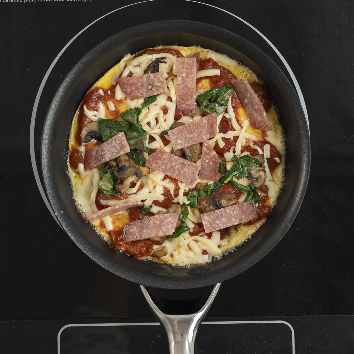 Egg in fry pan topped with tomato paste, cooked mushroom and spinach, cheese and sliced salami.