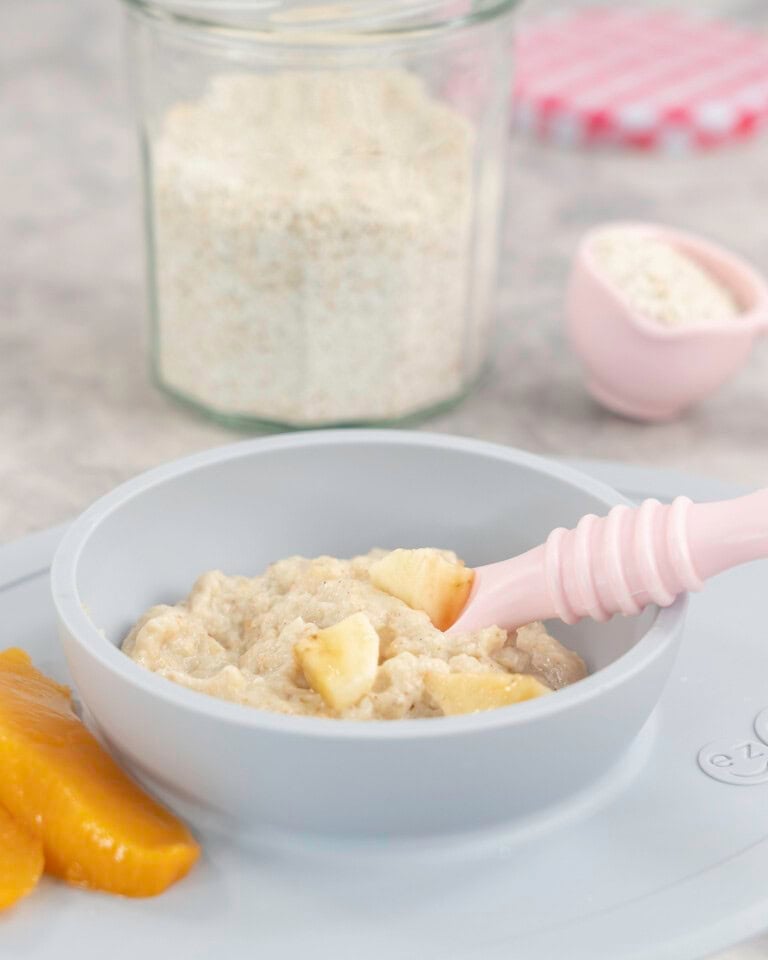 Silicone baby bowl and placemat on marble bench top with baby oats inside, topped with chopped banana pieces with baby spoon inside and two slices of canned peaches on the side, jar of ground oats on benchtop.