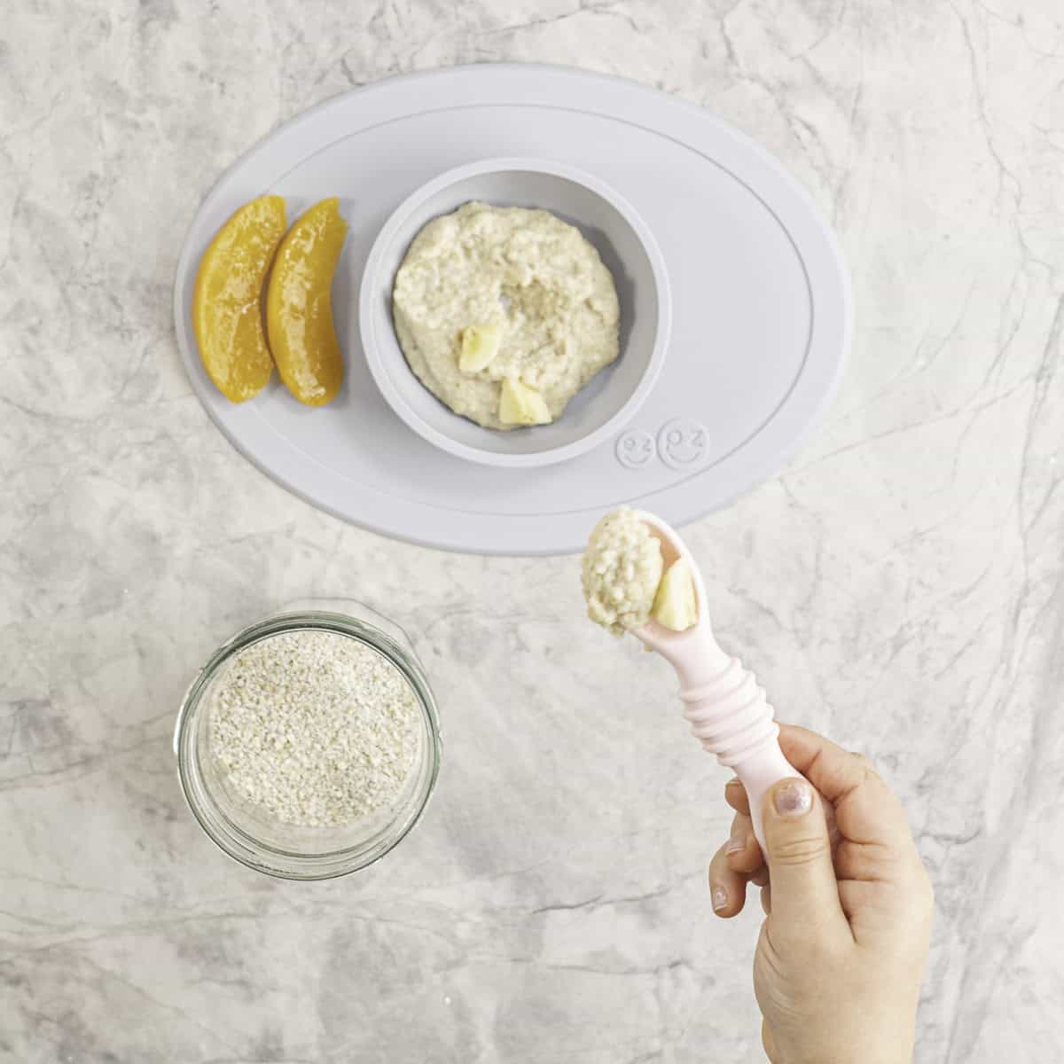 Toddler hand holding a silicone pink baby spoon with oatmeal and banana on its baby placemat and bowl with oats, and sliced canned peaches below next to a jar of ground oats on benchtop.