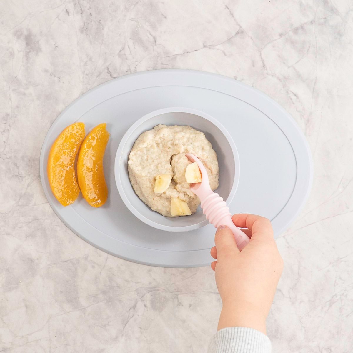 Silicone baby bowl and placemat on marble bench top with baby oats inside, topped with chopped banana pieces with toddler holding a baby spoon scooping onto spoon and two slices of canned peaches on the side.