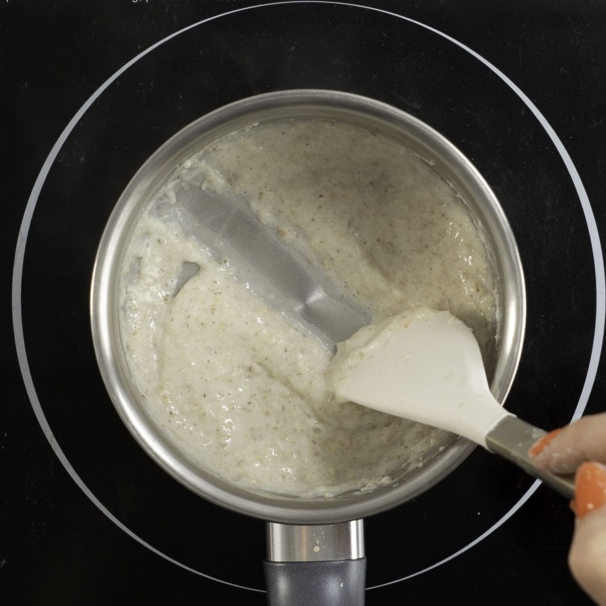 Small pot sitting on an electric stove top with creamy oats inside, hand sliding a spatula through oats.