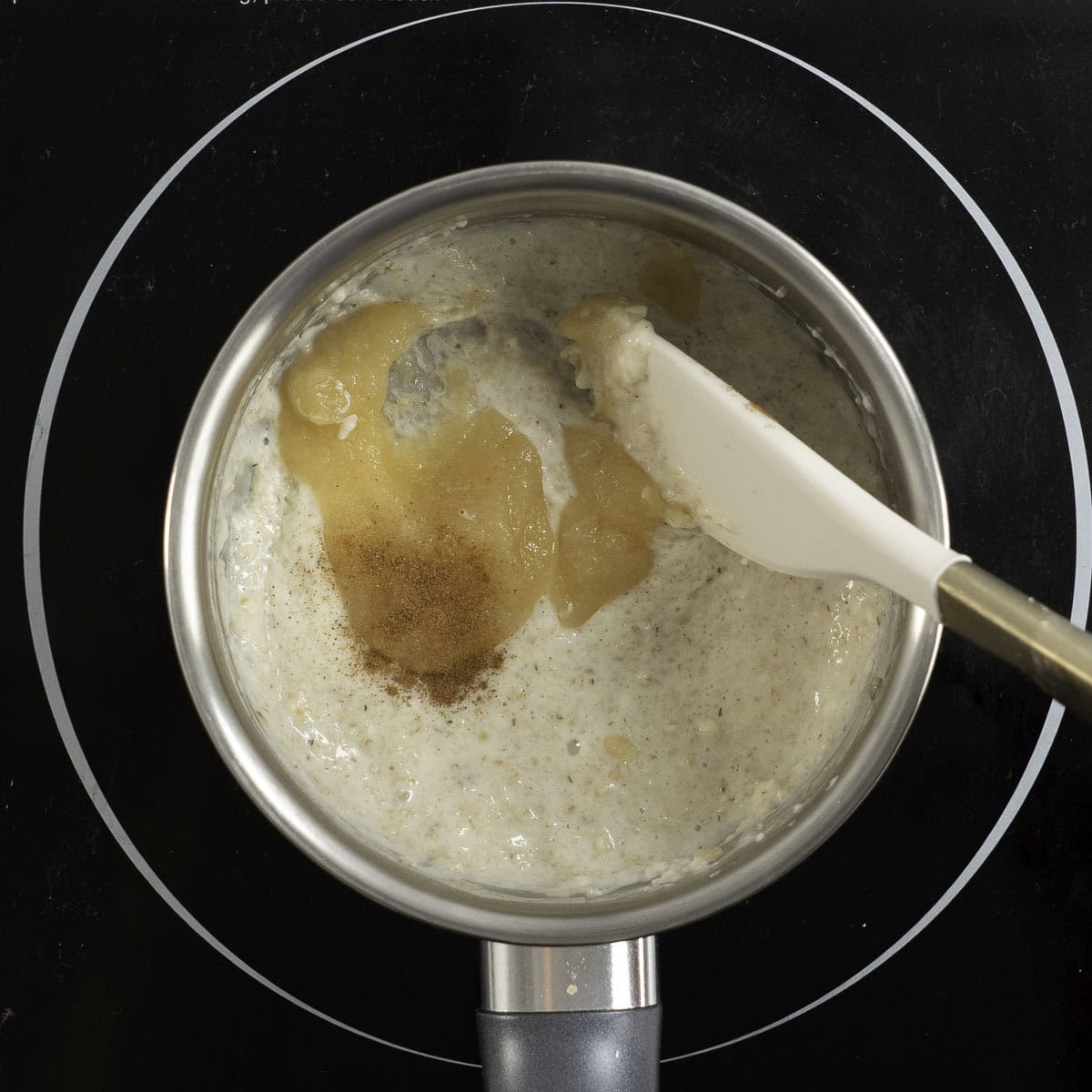 Small pot sitting on an electric stove top with creamy oats inside, apple puree and cinnamon added in with spatula stirring.