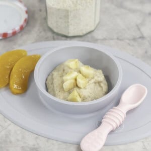 Silicone baby bowl and placemat on marble bench top with baby oats inside, topped with chopped banana pieces and two slices of canned peaches on the side. Pink baby spoon and jar of ground oats on benchtop.
